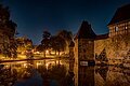 Die Weißenburger Stadtmauer am Abend. Das Gebäude spiegelt sich im Seeweiher und aus der Stadt scheinen Lichter, die dem Bild eine passende Atmosphere verleihen.