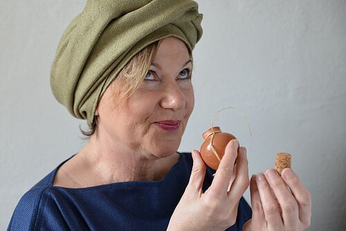 Eine Frau mit grünem Turban riecht an einem kleinen runden Tongefäß.