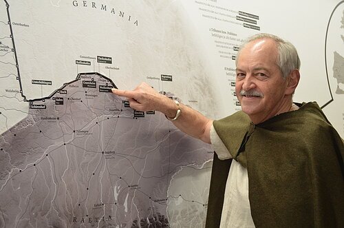 Ein Mann mit einem grünen altertümlichen Umgang schaut lächelnd in die Kamera und zeigt dabei mit seinem Zeigefinger auf eine Landkarte.