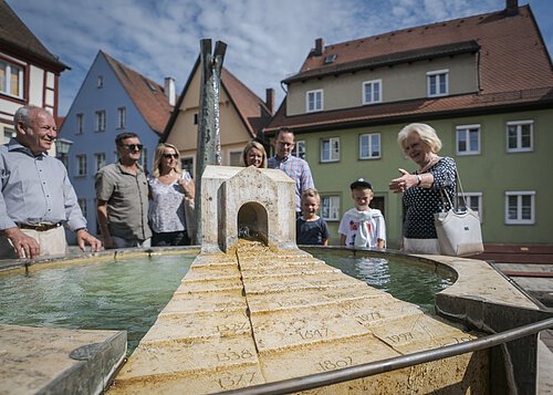 Eine Gruppe von Touristen steht um den Milleniumsbrunnen und blickt einen ältere Dame an die spricht. In der Mitte des kleinen brunnen steht ein kleines Haus aus Stein. Im Hintergrund stehen Häuser.