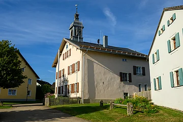 Zwei Gebäude mit Fensterläden und einem Turm, umgeben von einem Garten und einem Baum.