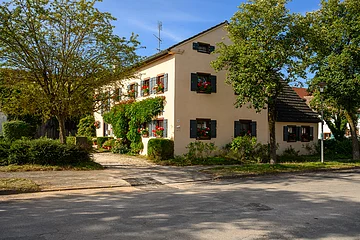 Das Familienhaus ist einen hellen Ton angestrichen und hat grüne Fensterläden, an den Fenstern befinden sich Blumenkästen mit Geranien. Um das Haus befinden sich verschiedene Sträucher und an der westlichen Häuserfront befinden sich Pflanzen