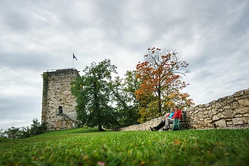 Zwei Personen sitzen auf einer Bank neben einer Steinmauer vor einem Turm und herbstlich gefärbten Bäumen.