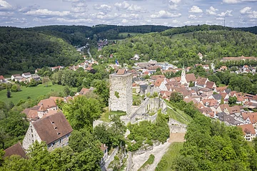 Luftaufnahme einer Burg mit Turm und umliegendem Dorf in einer grünen Hügellandschaft unter bewölktem Himmel.