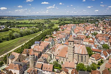 Luftaufnahme einer Stadt mit roten Dächern, zwei Türmen und einer großen Kirche, im Hintergrund grüne Felder und ein See.