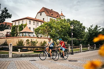 Zwei Personen mit Fahrradhelmen fahren auf einem gepflasterten Weg vor einem historischen Gebäude mit Bäumen.