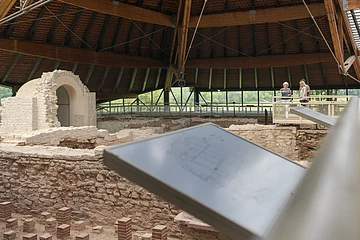 Überdachte archäologische Ausgrabung mit zwei Personen auf einem Holzsteg und Informationstafel im Vordergrund.