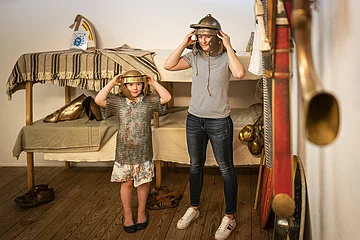 Kind und Frau stehen in einem Raum mit historischer Rüstung, beide tragen Helme und schauen in die Kamera.