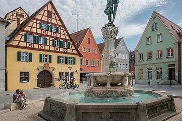 Brunnen mit Statue auf einem Platz, umgeben von Fachwerkhäusern und Menschen, unter bewölktem Himmel.
