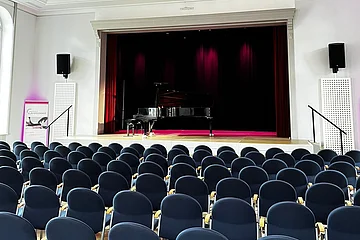 Leerer Konzertsaal mit Reihen blauer Stühle und einem Flügel auf der Bühne unter einer Deckenleuchte.