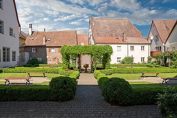 Innenhof mit gepflegtem Garten, Bänken, Hecken und alten Häusern mit roten Ziegeldächern im Hintergrund.