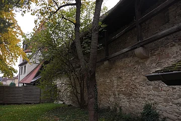 Steinmauer mit Holzbalken, dahinter Bäume und ein Haus mit rotem Dach im Herbst.